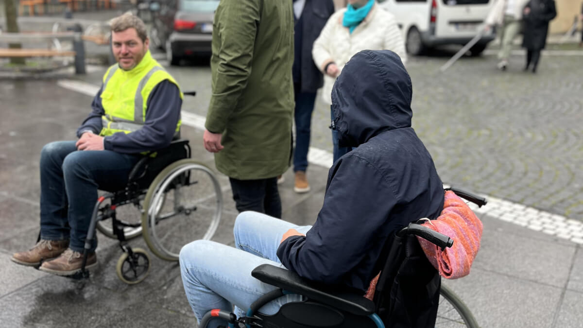 Dans une rue pavée, deux personnes en fauteuil roulant au milieu d'un groupe de piétons.
