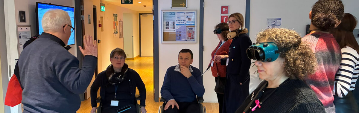 Dans un couloir, des participants testent des lunettes de simulation de déficience visuelle et des fauteuils roulants, guidés par un animateur.